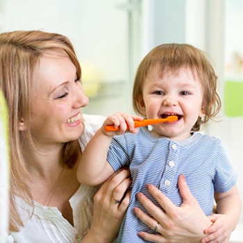 A mother smiling while her baby brushes their teeth using a manual toothbrush