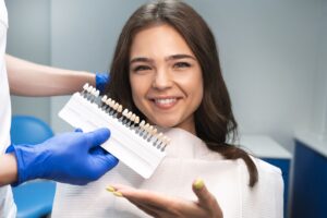 Woman with brown hair in dental chair with white bib while dentist holds veneer samples up to her face